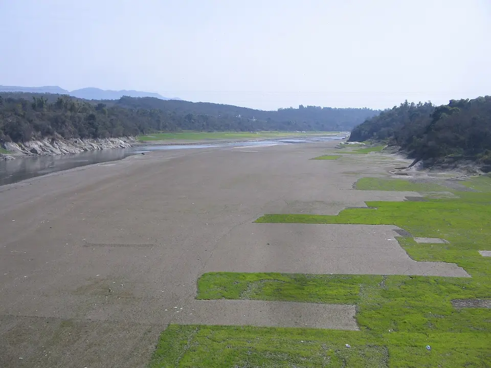 A wide, mostly dry riverbed at Amb, with strips of green field and low hills behind